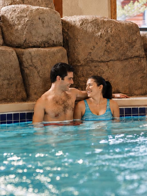 A man and a woman smile at each other while relaxing in a pool, surrounded by large, textured stone blocks.