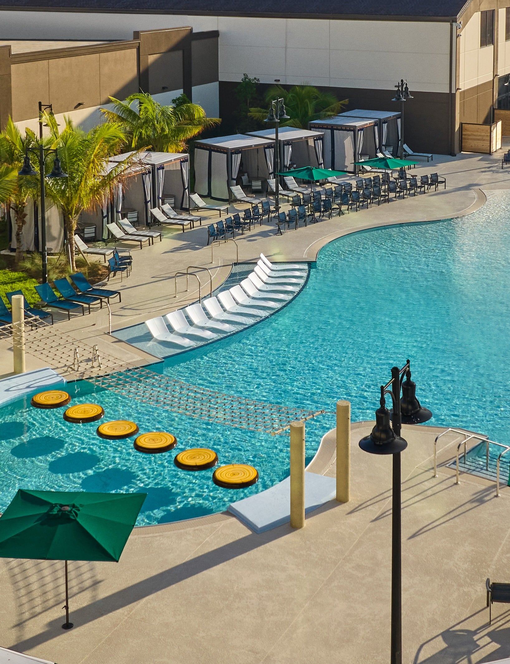 A resort pool area with lounge chairs, cabanas, a net crossing over water, and circular floatables. Palm trees and umbrellas provide shade.