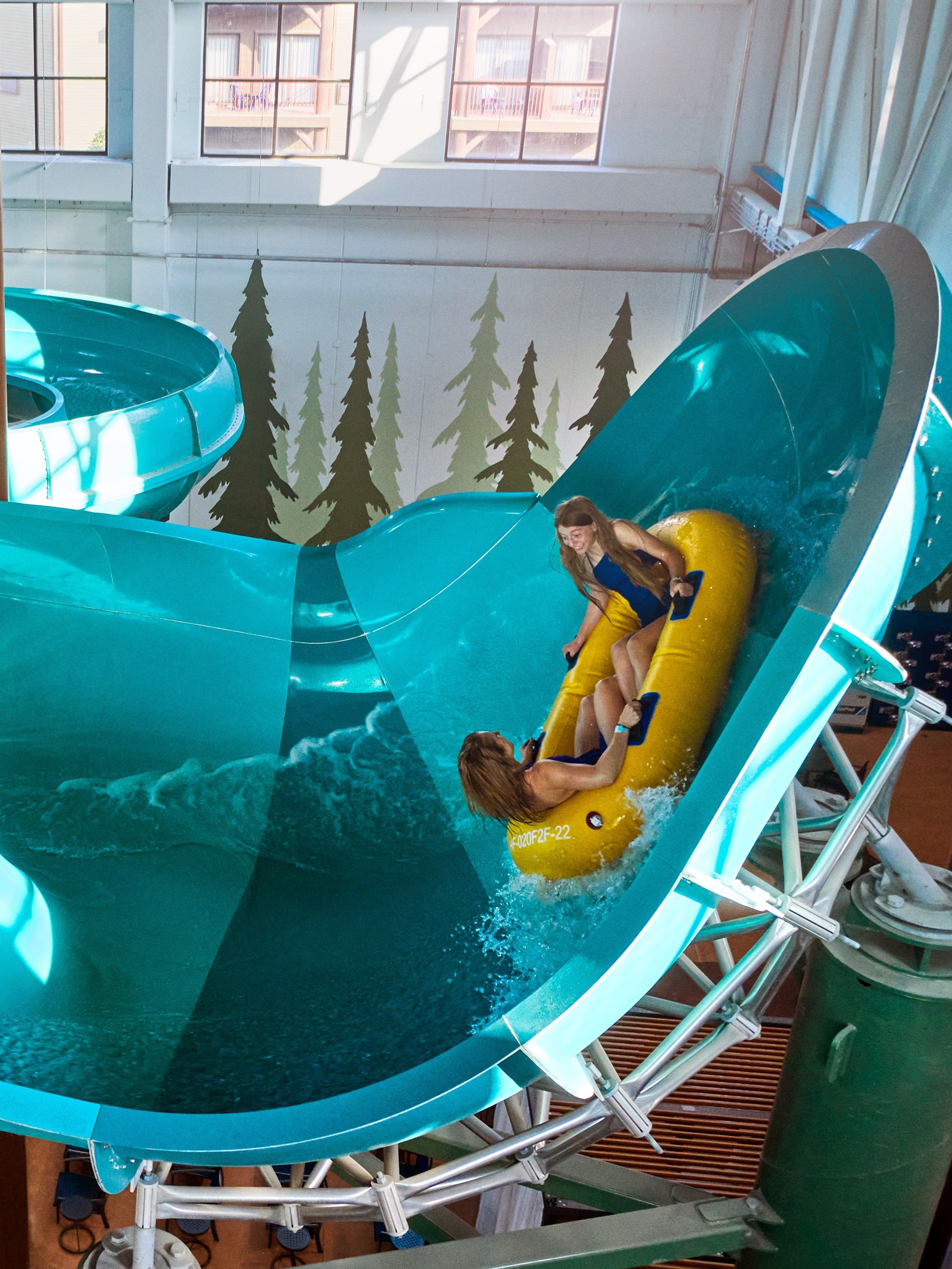 Two people enjoy a ride on a yellow inflatable raft down a large, blue indoor waterslide, with forest-themed decor on the walls.