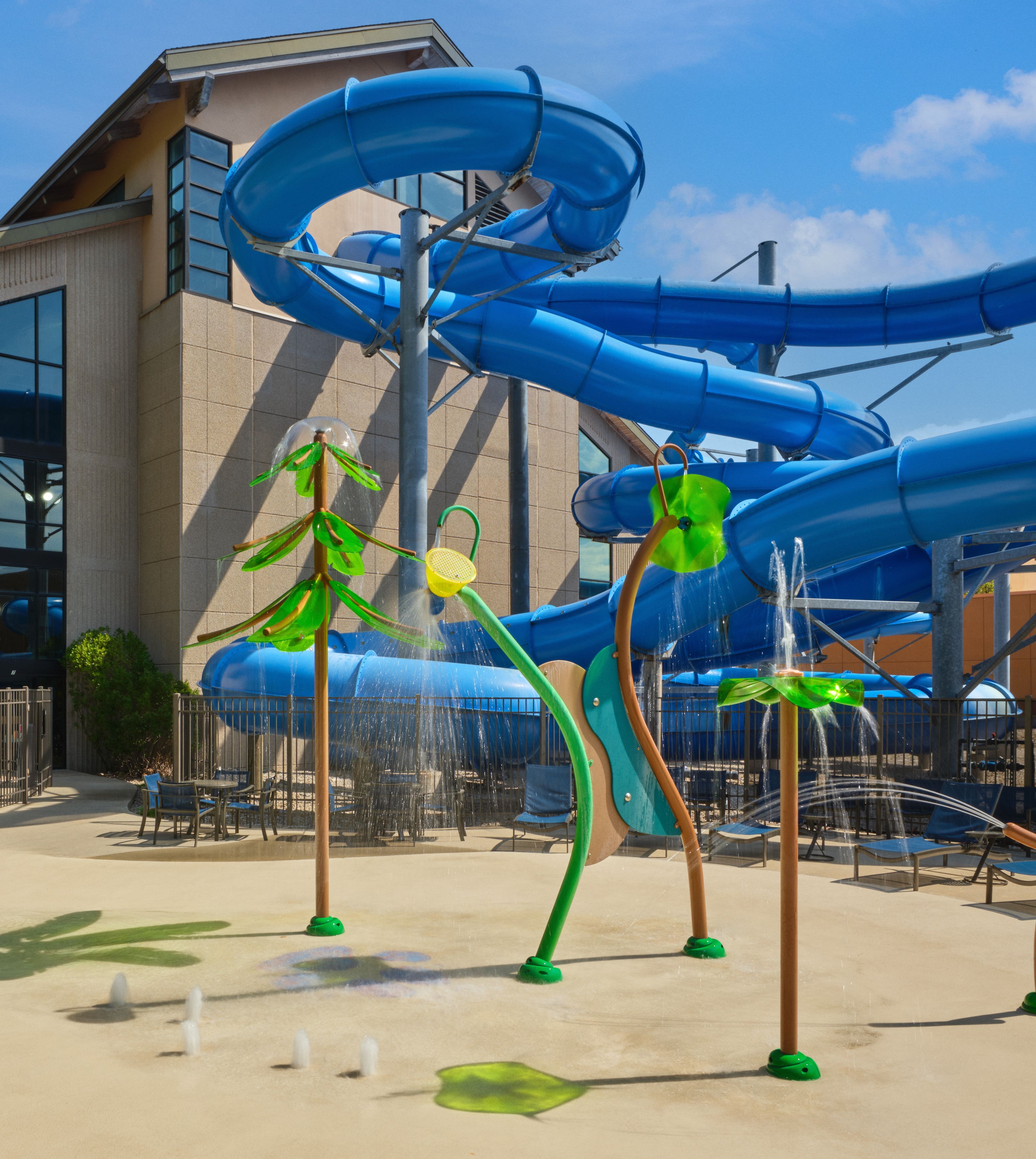 Water park with large blue slide, splash features shaped like trees, leafy sprayers, and a sunny sky in the background.