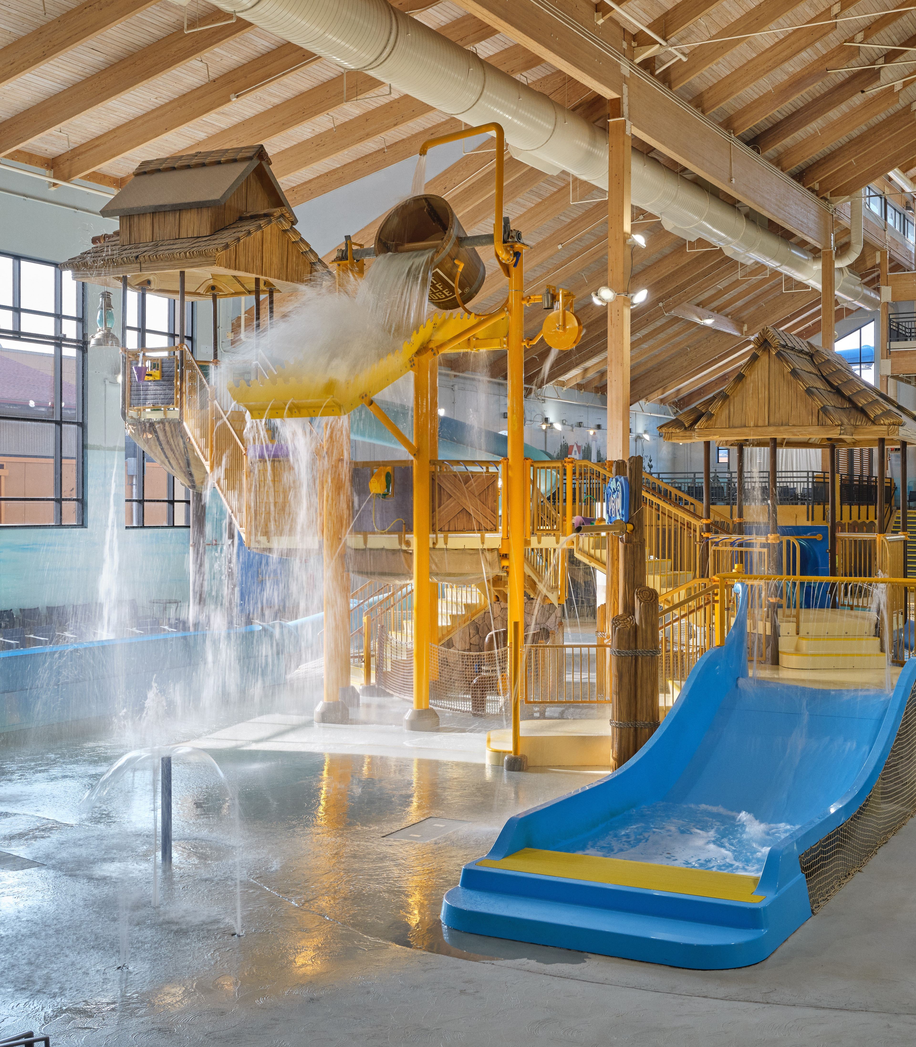 Indoor water park with slides, a large bucket dumping water, and splash zones under a wooden ceiling.