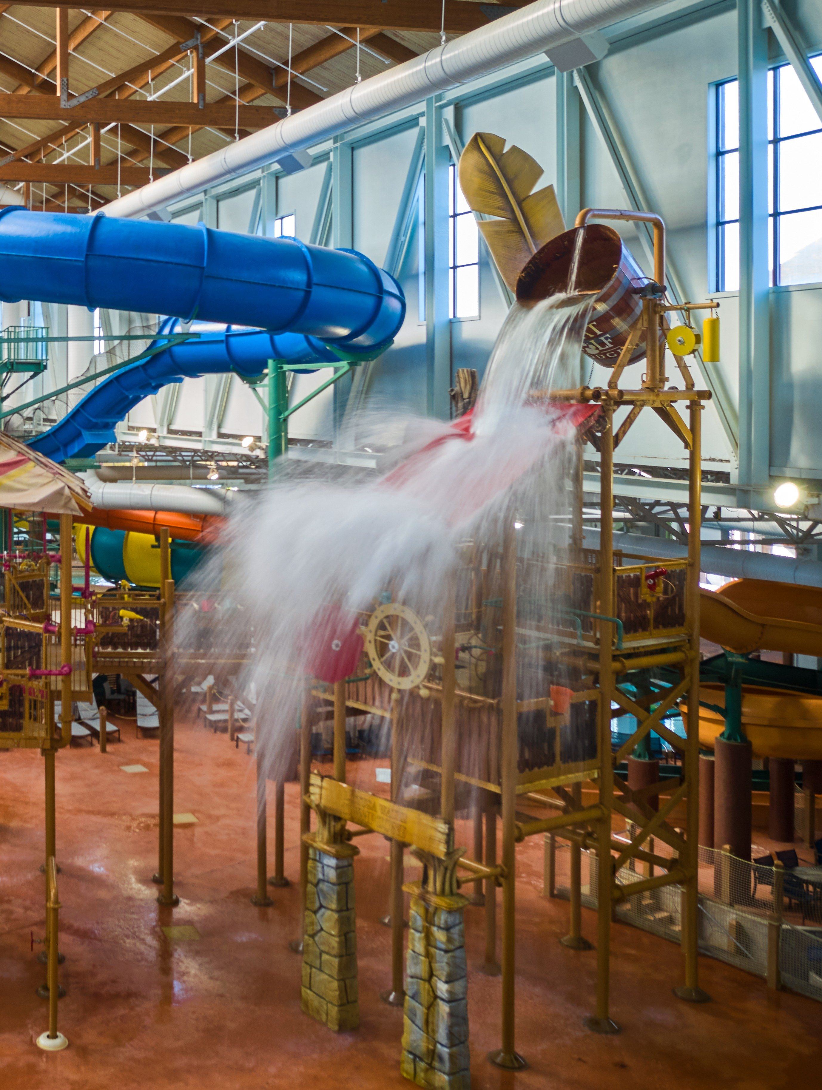 Indoor water park with large bucket dumping water onto a play structure, surrounded by slides and colorful pipes.