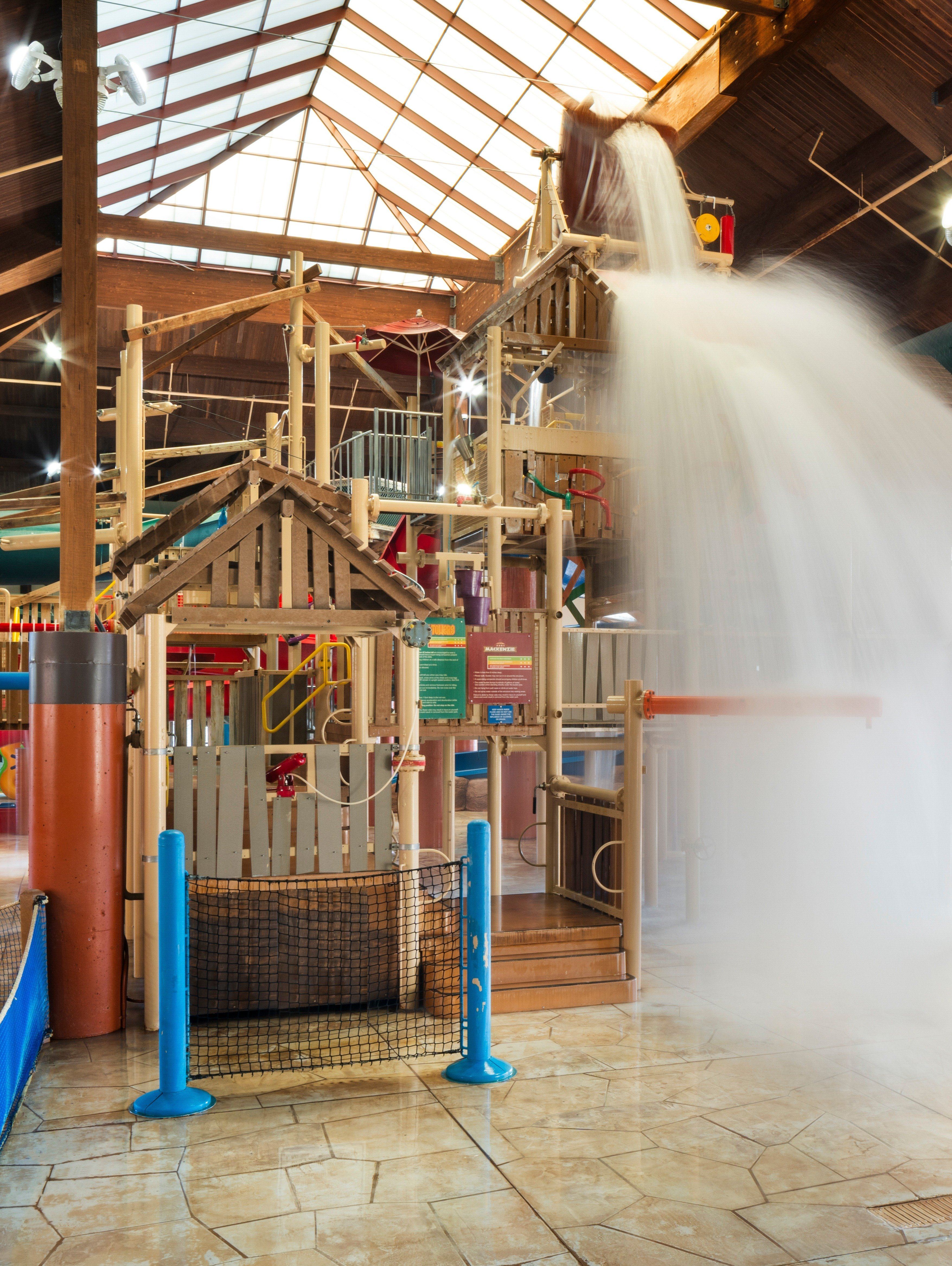Indoor water park with wooden play structures and a large bucket pouring water, under a skylit roof.