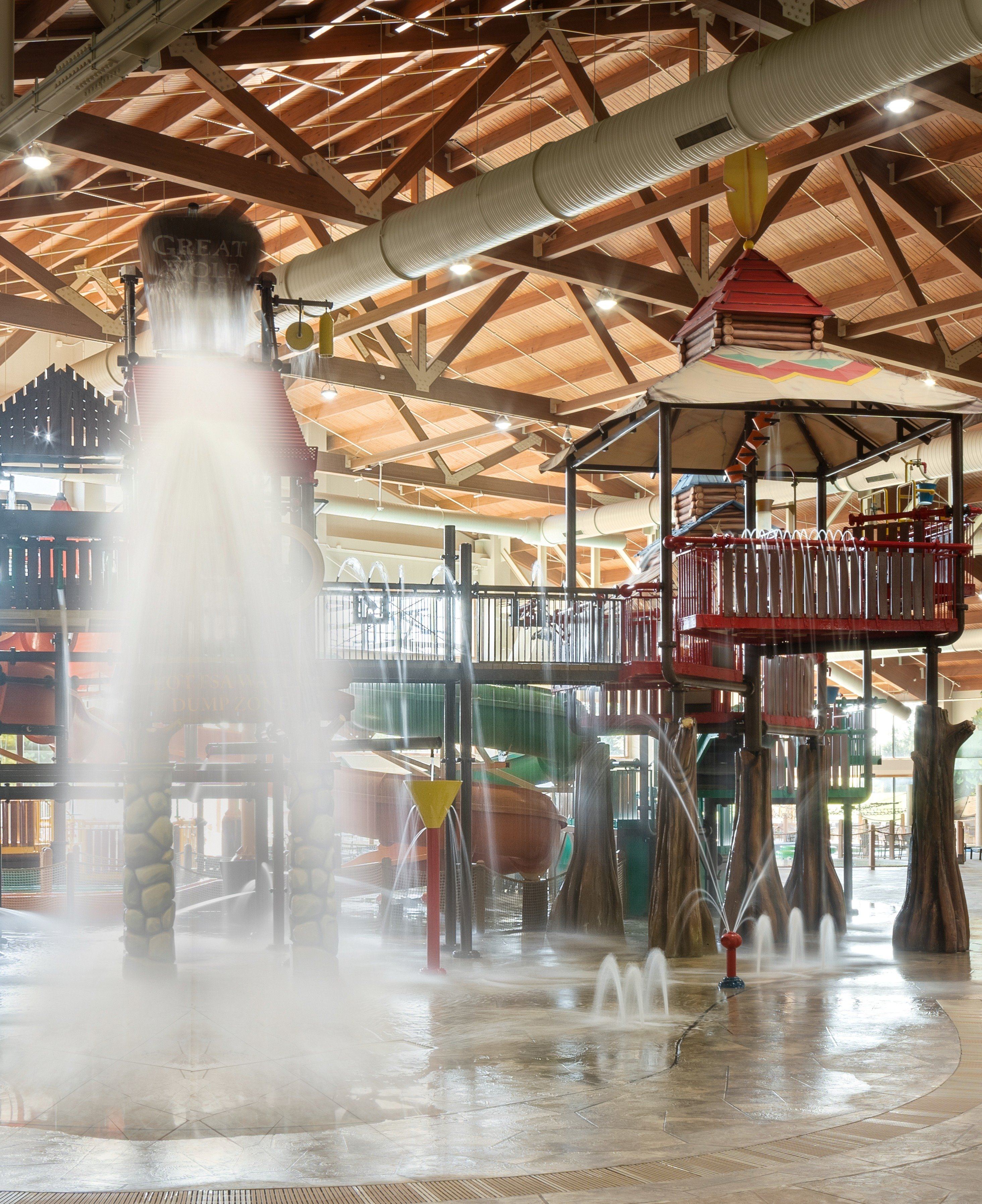 Indoor water park with wooden beams, large water bucket pouring out, slides, and sprayers creating a playful atmosphere.