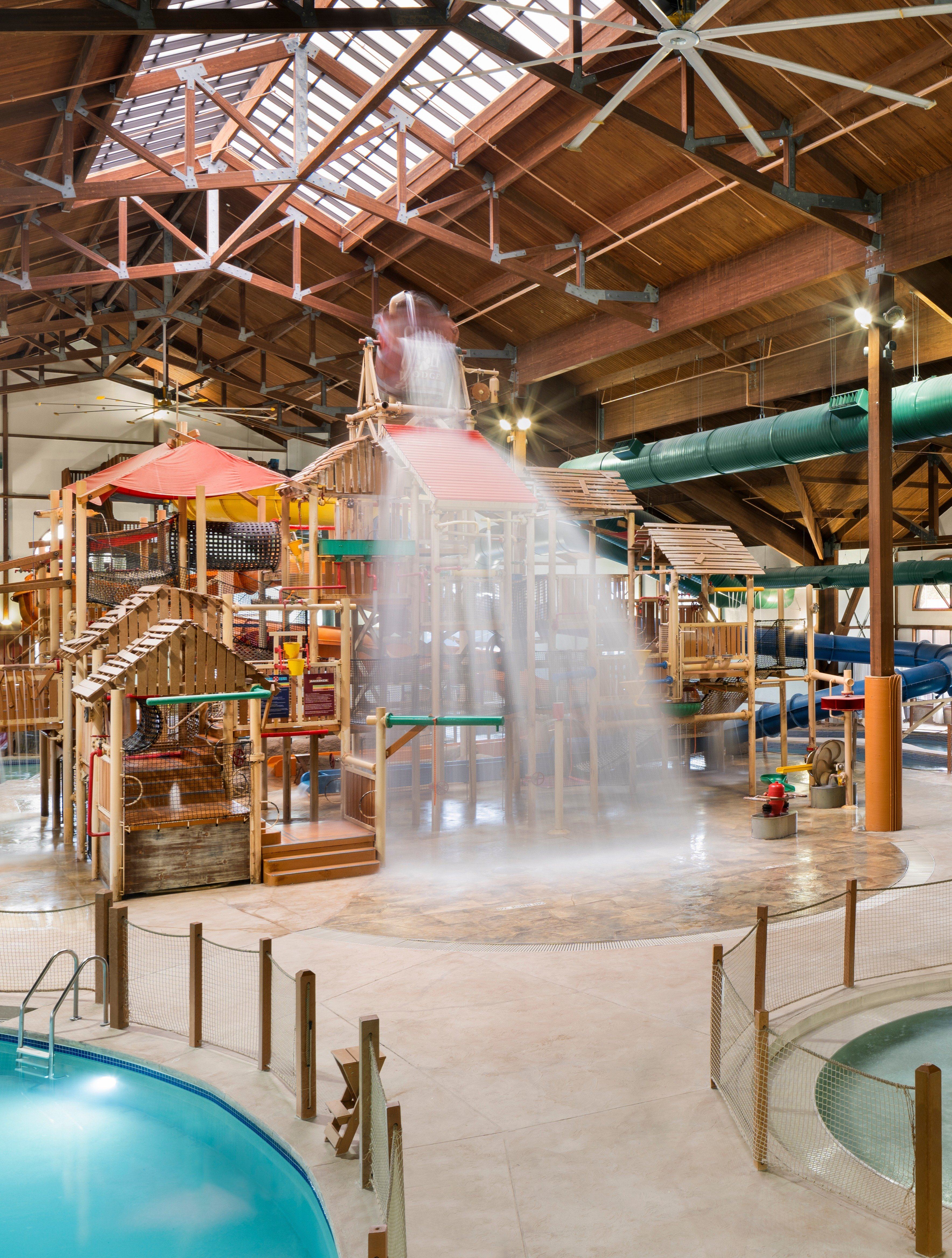 Indoor water park with a wooden play structure, water slides, and a large bucket pouring water. The space has a high ceiling and natural lighting.