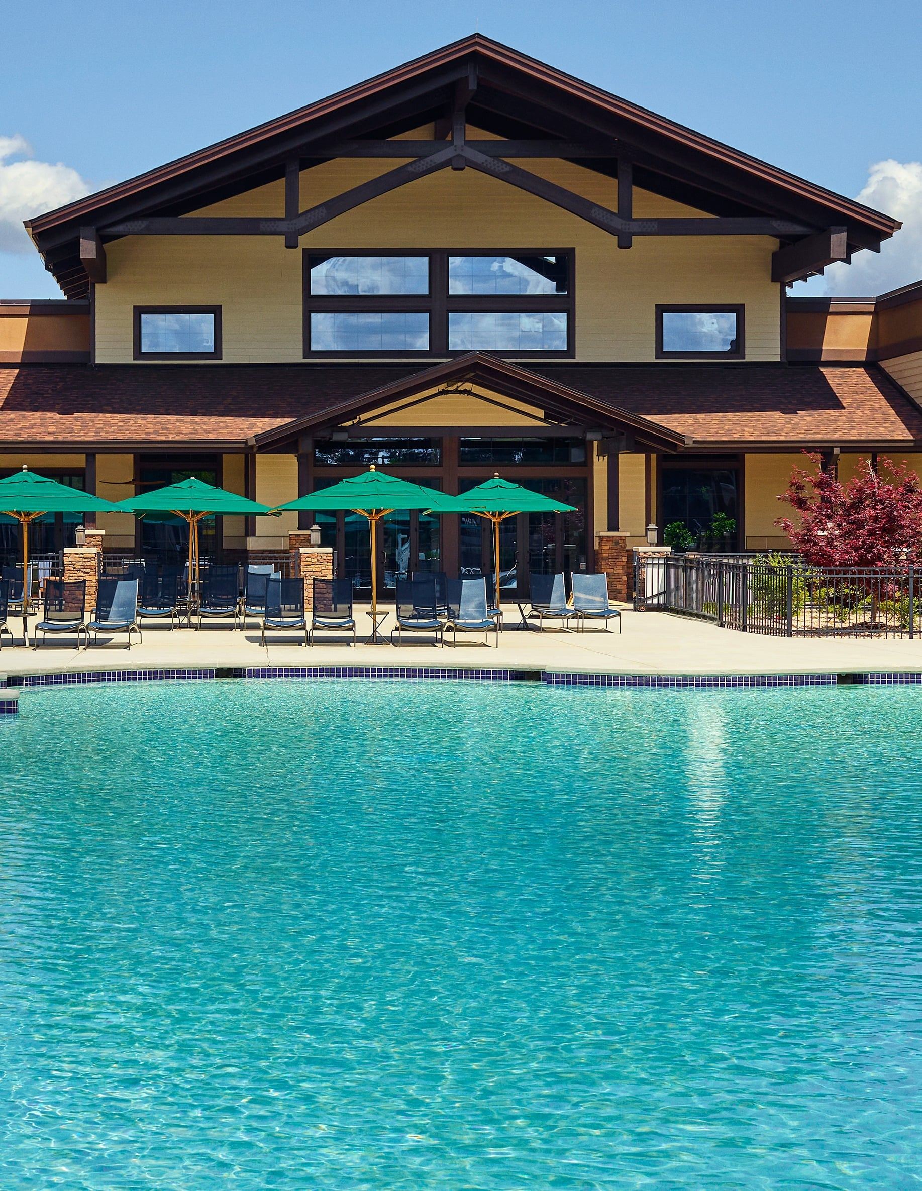 Large swimming pool in front of a modern building with green umbrellas and lounge chairs on the deck.