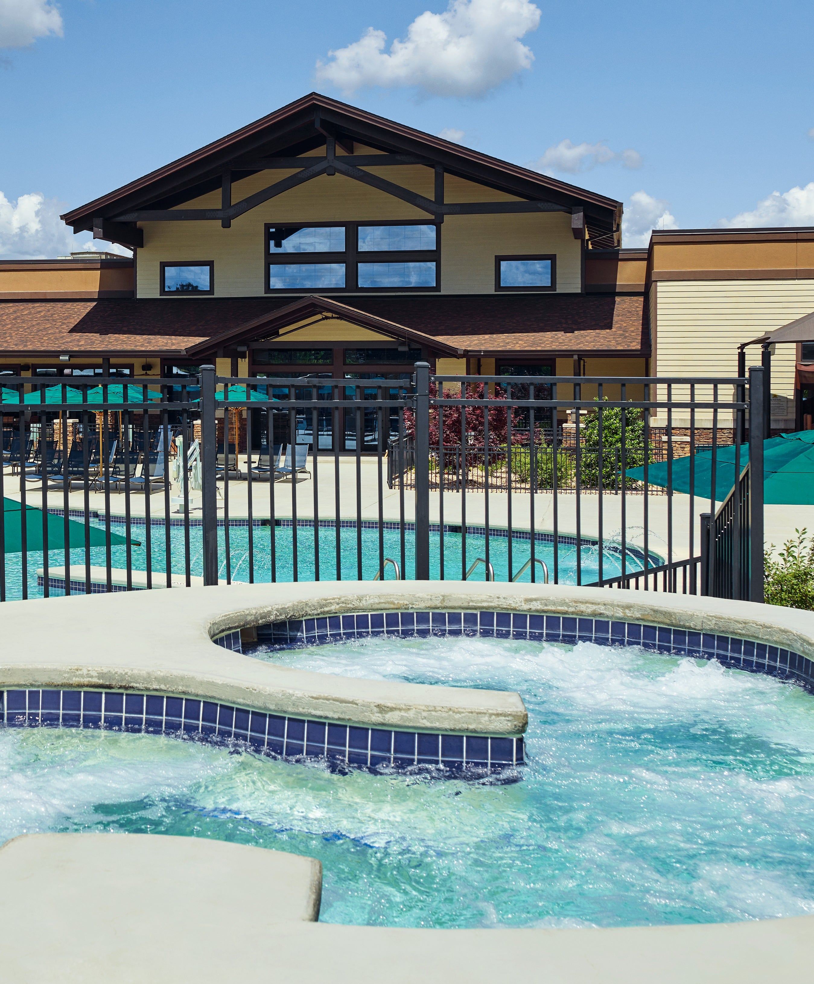 Outdoor hot tub with bubbling water, adjacent to a pool and fenced area, in front of a large building under a blue sky with clouds.