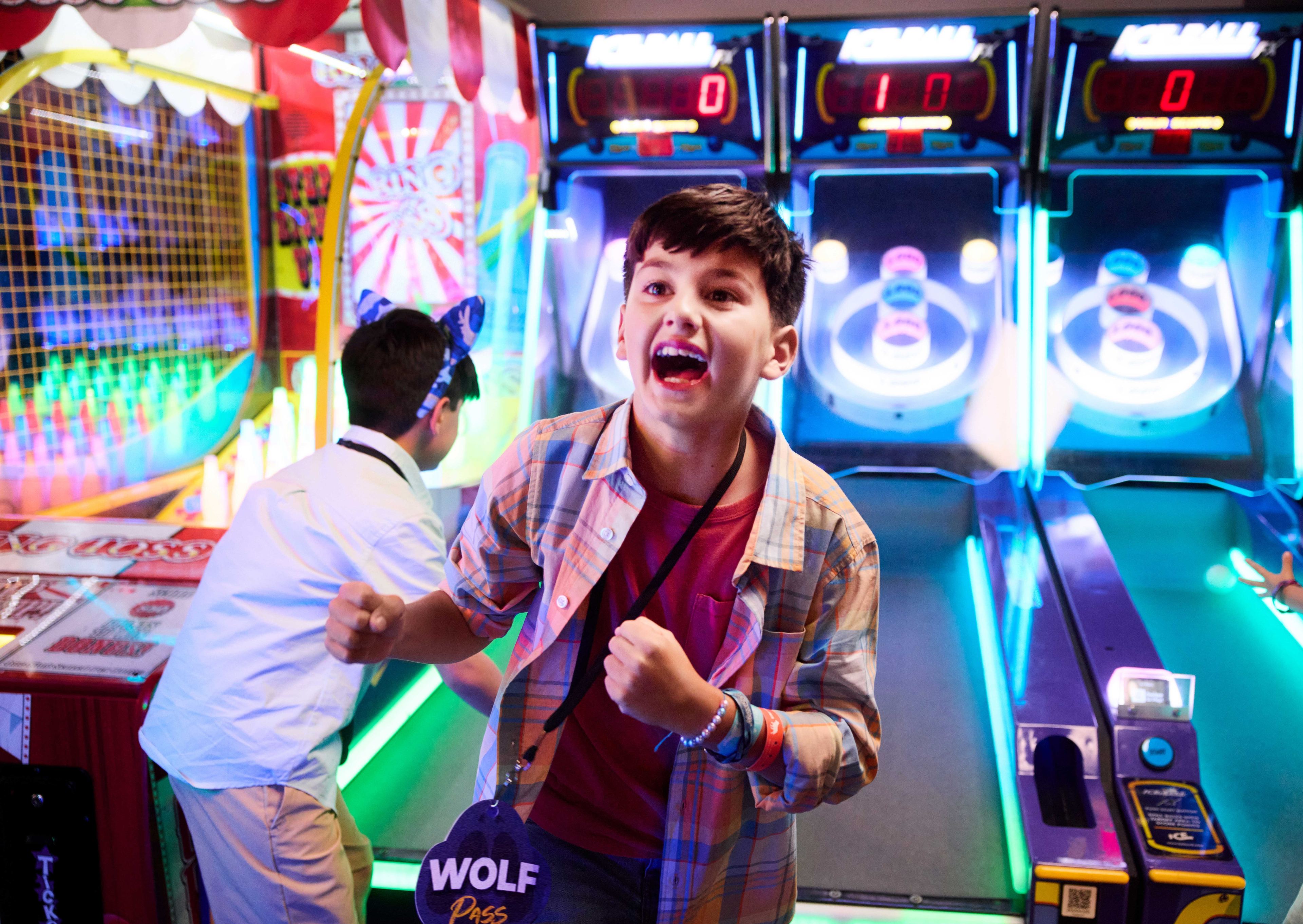 Excited child celebrates at an arcade, surrounded by colorful lights and skee-ball machines displaying scores in the background.