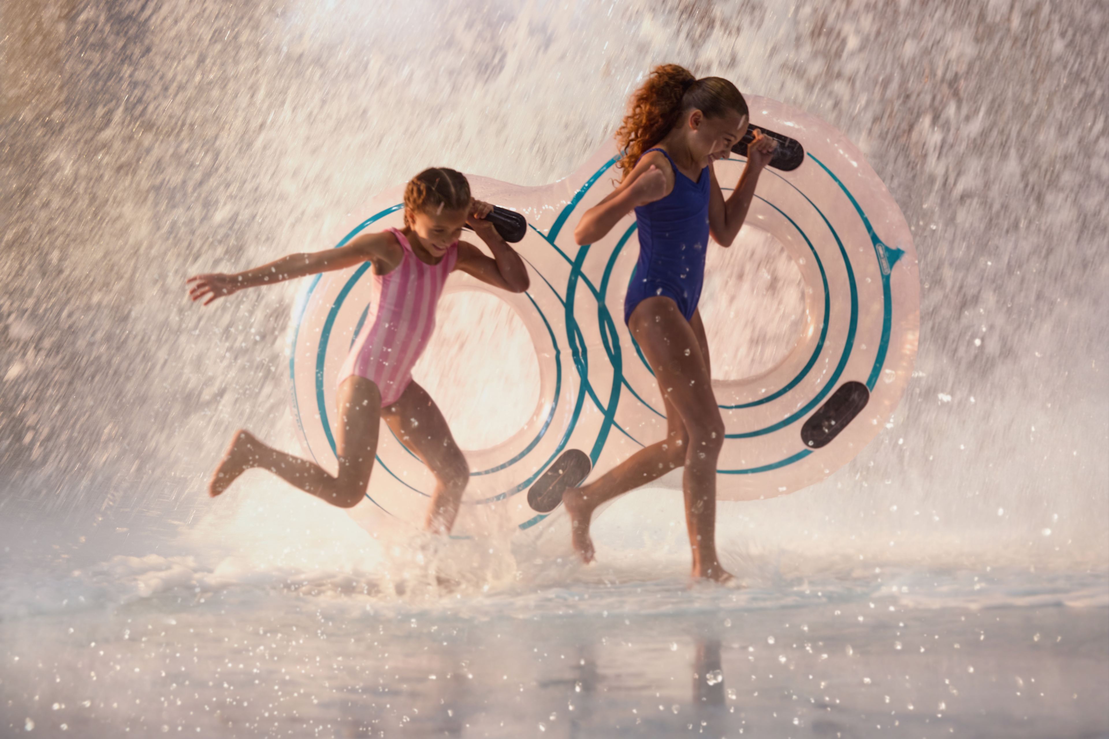 Two girls in swimsuits holding inflatable rings run through a curtain of water with playful expressions, splashing in a sunlit scene.