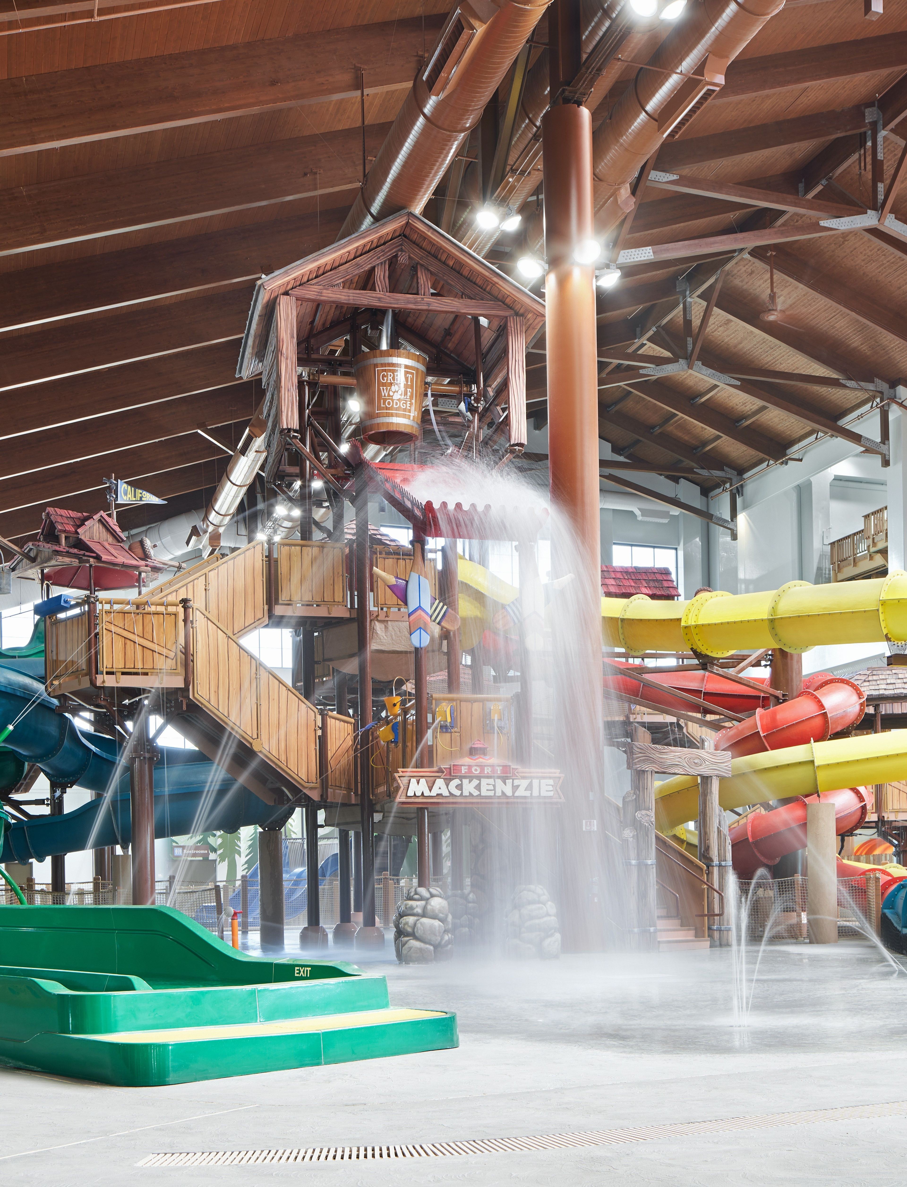 Indoor water park with colorful slides, wooden structures, and a large bucket pouring water over a play area.